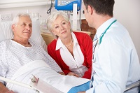 Image of a woman sitting next to an elderly male patient in hospital bed speaking with a doctor